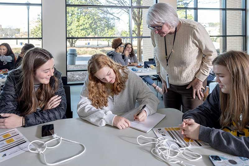 a class group around a table with a professor helping the students, with supportive communities for researchers across disciplines, promoting knowledge exchange, and enabling access to internal and external funding.